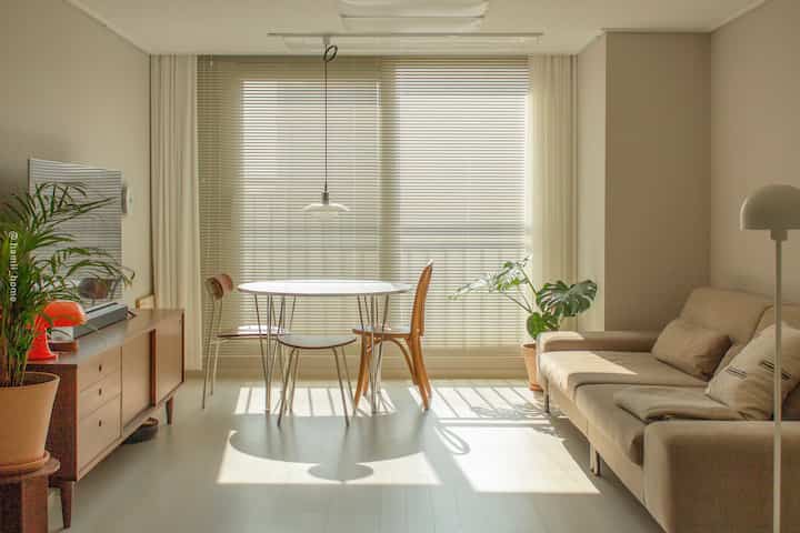 Natural tone living room with beige fabric sofa, round white table, wooden TV stand, and large window blinds creating a cozy atmosphere