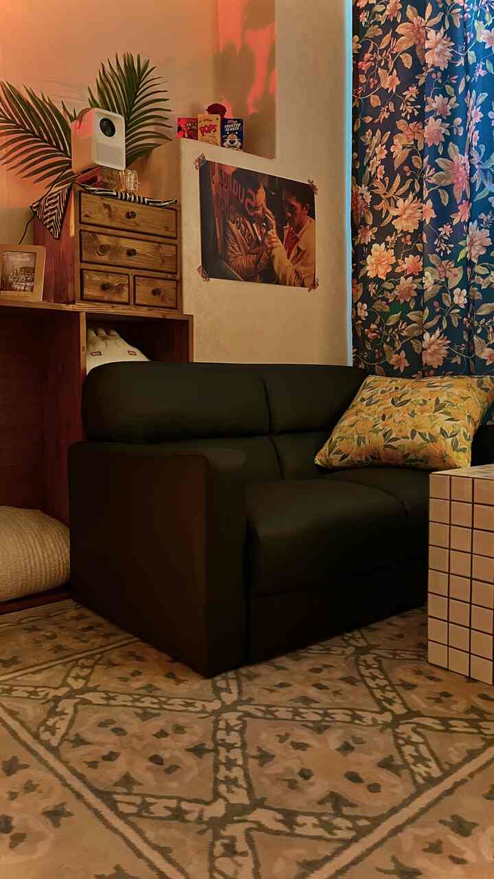 暖かいイエロートーンの小さなリビング、ブラックの2人掛けソファとヴィンテージラグ、レトロ映画ポスターとミニプロジェクターが配置された落ち着いた空間