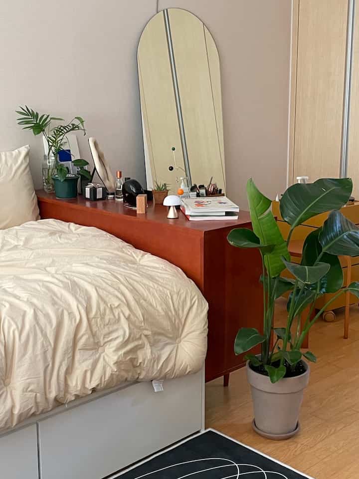 Natural-toned bedroom featuring beige bedding, reddish dresser, arched mirror, and large green plant creating a calm atmosphere