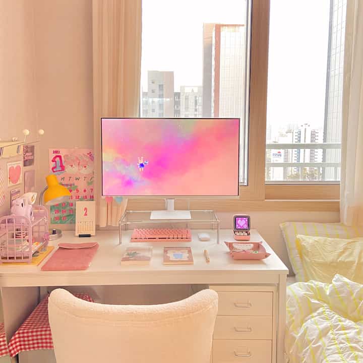パステル調のコンパクトな寝室兼ホームオフィス空間、ホワイトデスクとモニター、ふわふわの椅子、隣にベッドが配置された整ったインテリア