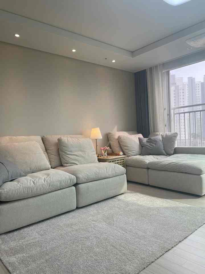 Beige toned living room with large sectional sofa and carpet, cozy and comfortable space by window with a lamp