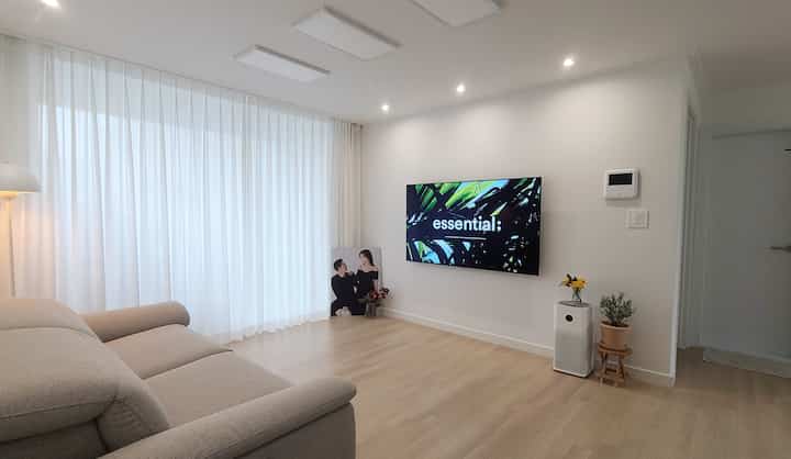 白とナチュラルカラーの調和がとれたリビング、ベージュの3人用リクライニングソファと大きな薄手の白いカーテンが配置された、清潔で広々とした雰囲気のインテリア。