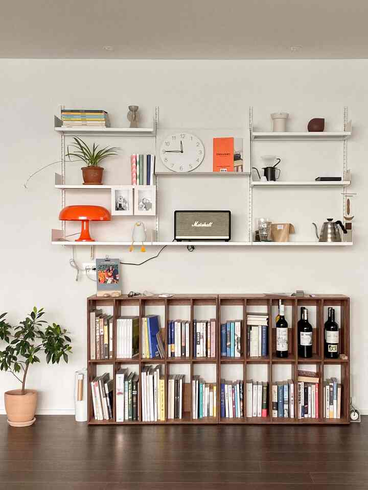 White walls and wood-tone bookshelves in a living room cum home office space, accented with an orange lamp and plant on shelves, cozy atmosphere