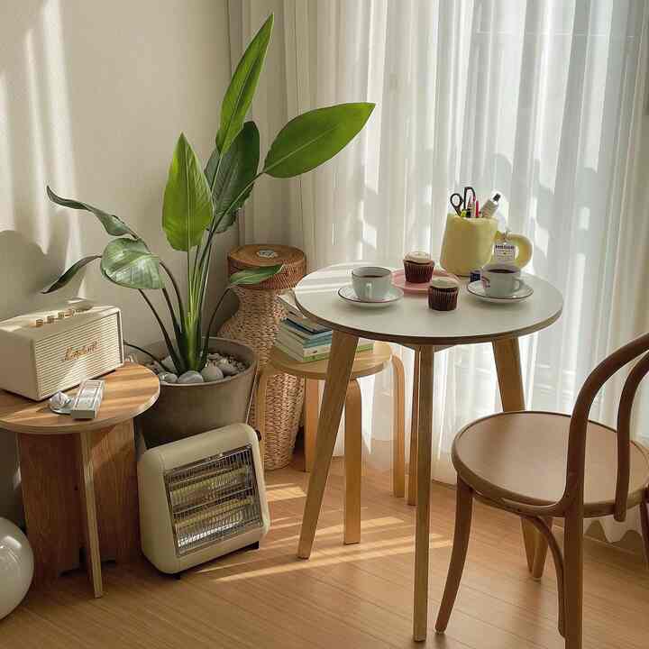 白と木目調の落ち着いたワンルームのホームカフェ空間、丸テーブルと木製椅子、大きな観葉植物と小型ヒーターが配置された雰囲気のあるインテリア