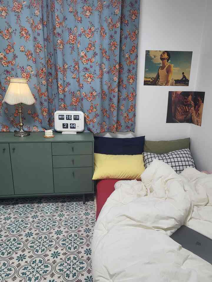 White and blue toned bedroom featuring vintage floral curtains, green nightstand, and colorful pillows arranged on the bed in a cozy space