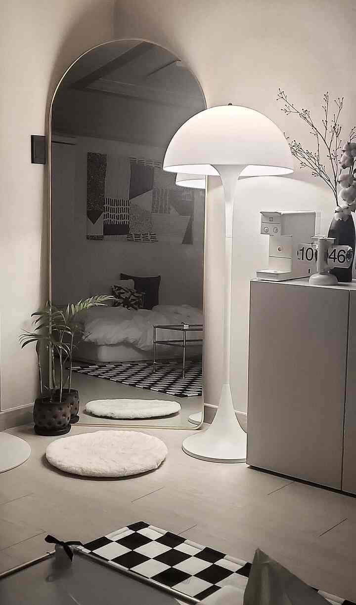 白とモノトーンでまとめられたワンルーム寝室、大きなアーチ型の鏡とフロアランプ、植物が配置された落ち着いた印象的な空間