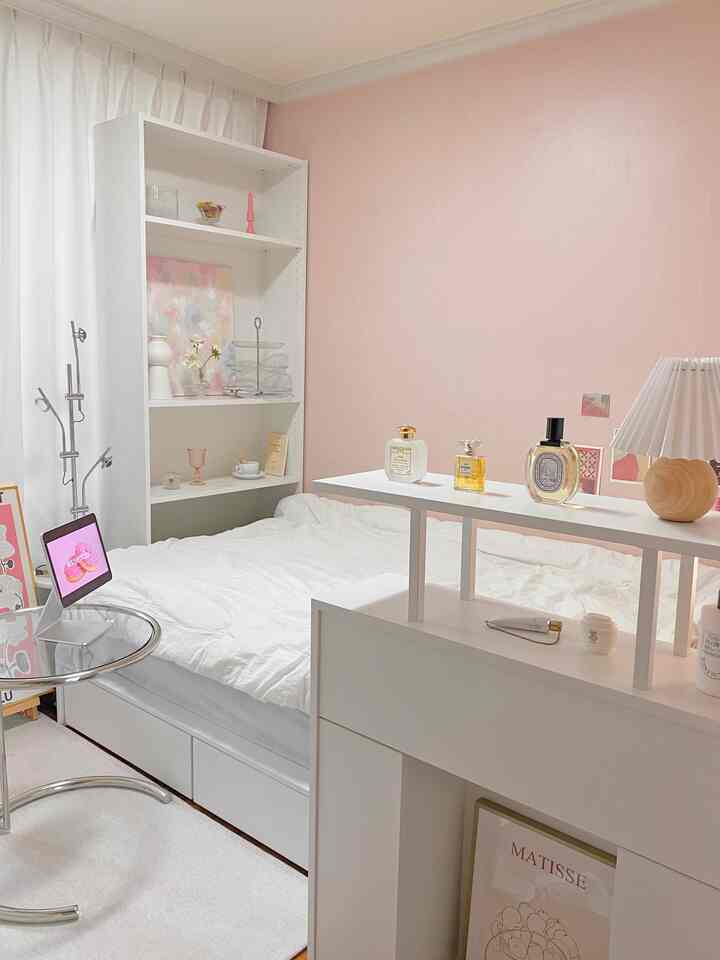 A cozy studio bedroom with pink walls and white furniture, featuring a console table with perfume and lamp, along with a bookshelf