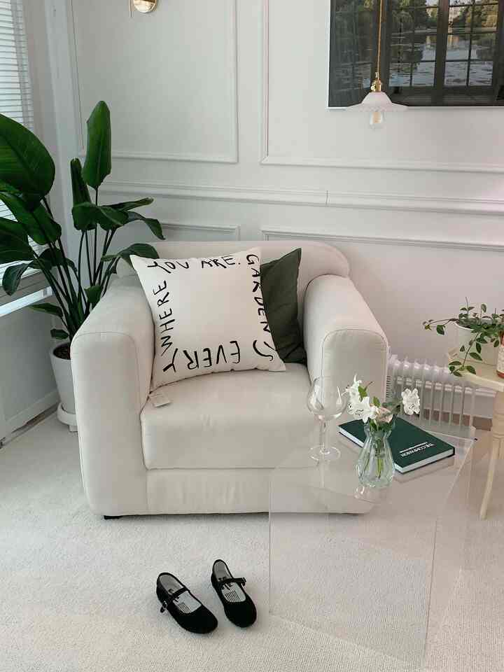 White and natural toned living room featuring beige armchair, transparent acrylic table, and plants in a clean minimalist space