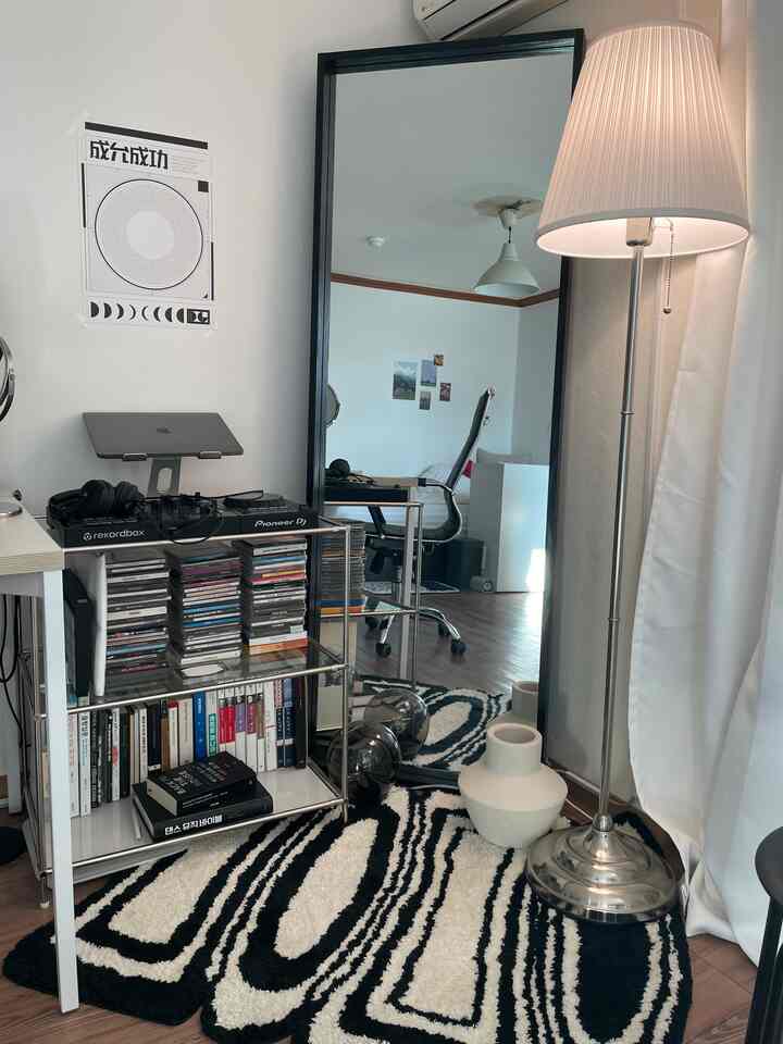 Modern living room with white and black patterned rug, featuring a bookshelf, full-length mirror, and floor lamp, evoking a remote work setup