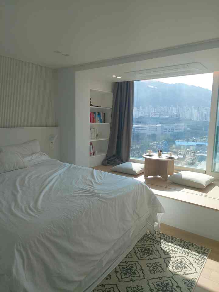 White and beige toned bedroom featuring large bed, window-seat area, and bookshelf with cozy, clean atmosphere