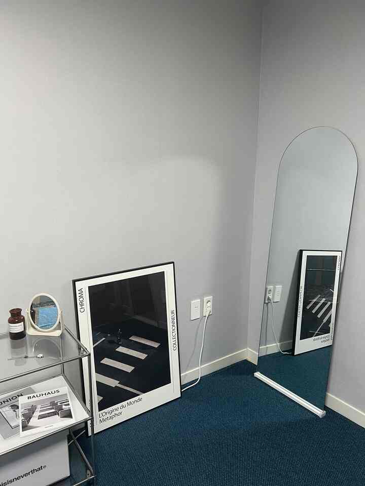 チャコールグレーの壁とブルーのカーペットが敷かれた小さな部屋、アーチ型の全身鏡とポスターが配置されたシンプルでモダンなインテリア