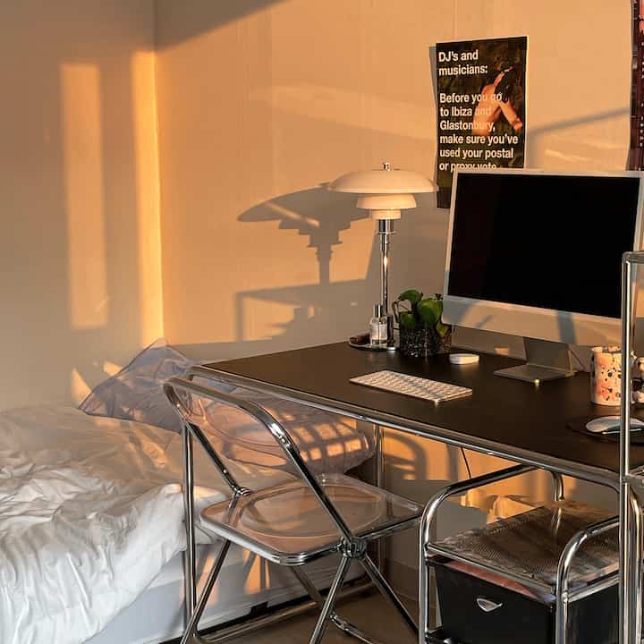 Warm beige-toned studio home office with morning sunlight, featuring a transparent folding chair, desk with monitor, and bed in harmony