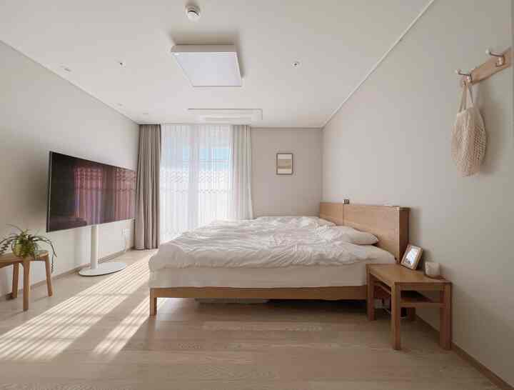 Bright bedroom in white and natural tones featuring a large bed, side table, TV stand, and sheer curtains creating a clean atmosphere