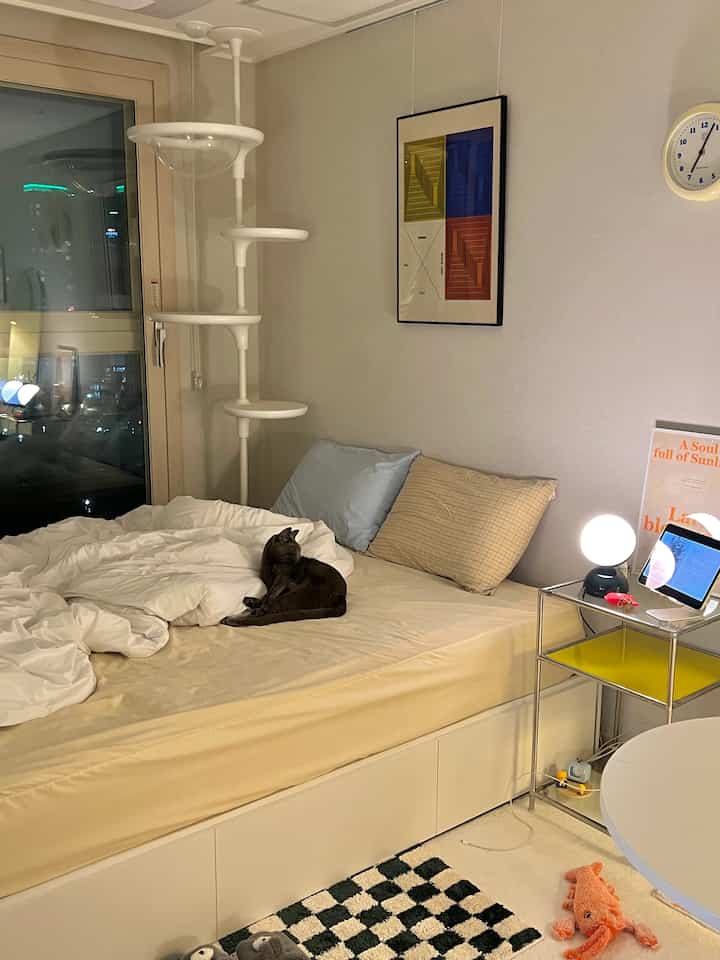 A cozy bedroom with soft beige and white tones featuring a black cat on the central bed, a white cat tower on the left, and stainless steel side table with round table lamp on the right