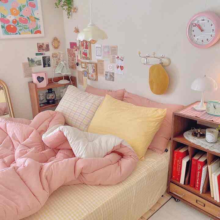 Cozy bedroom with pink and yellow tones, featuring a bed, colorful posters, storage shelf, and a wall clock creating a warm atmosphere