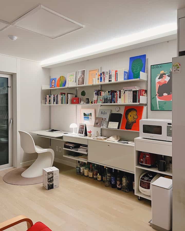 A white and wood-tone compact home office featuring wall-mounted bookshelves and artistic decor, creating a cozy atmosphere