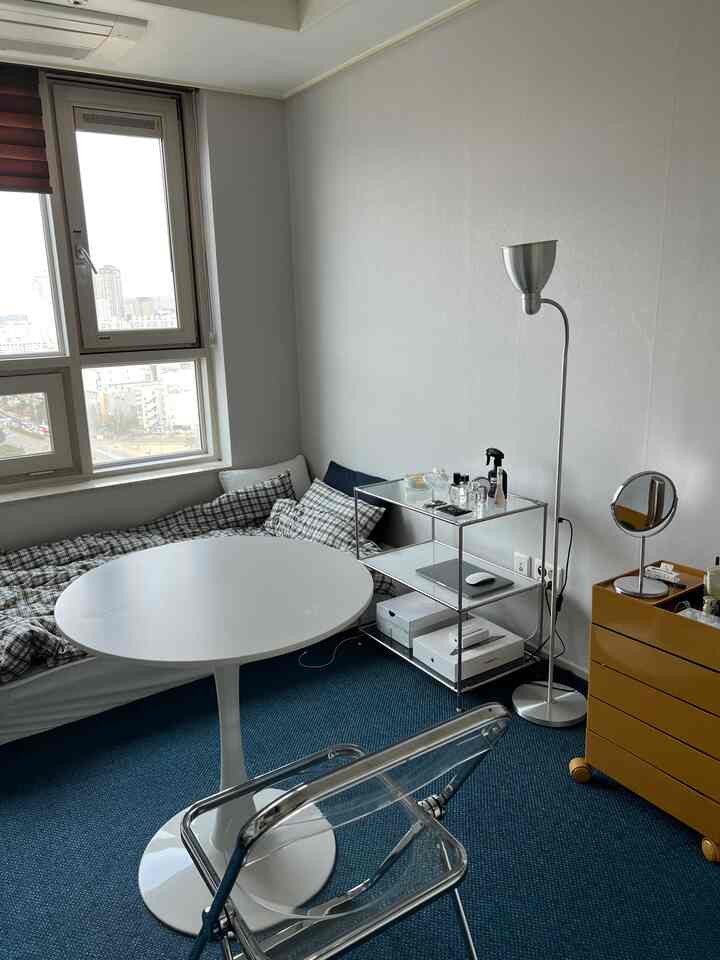 白い壁と青いカーペットのワンルーム、透明な折りたたみ椅子と丸テーブル、3段のモジュール棚とフロアランプが配置されたモダンな空間