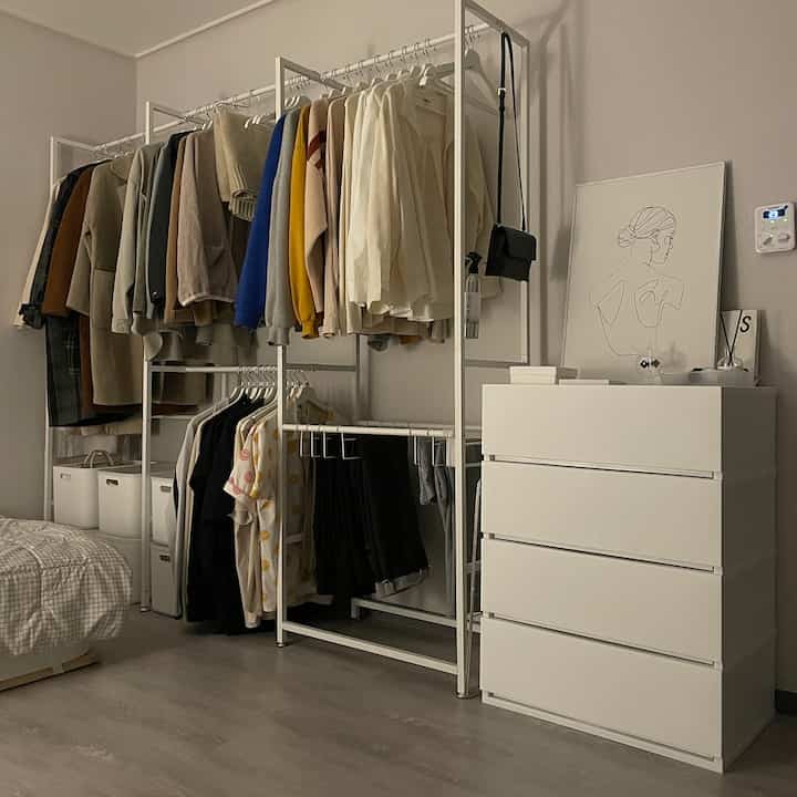 Simple minimalist bedroom featuring an open white closet rack and dresser with natural toned flooring