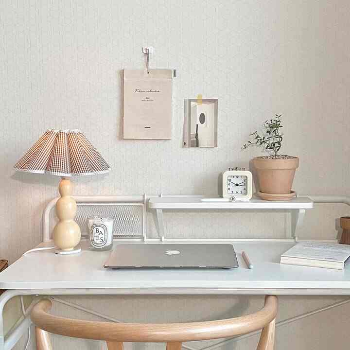 Minimal home office with white and beige tones featuring desk with laptop, candle, and table lamp creating a simple workspace