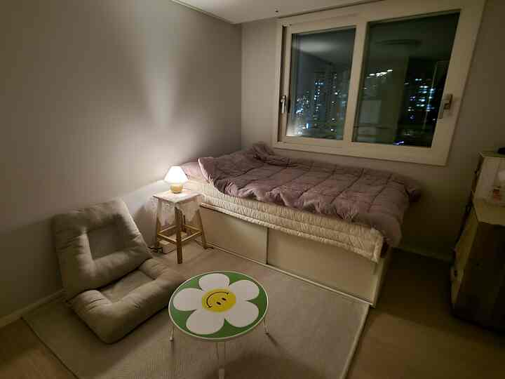 A compact bedroom in beige and white tones featuring a single bed, cushioned lounge chair, round table, and soft lighting creating a cozy atmosphere