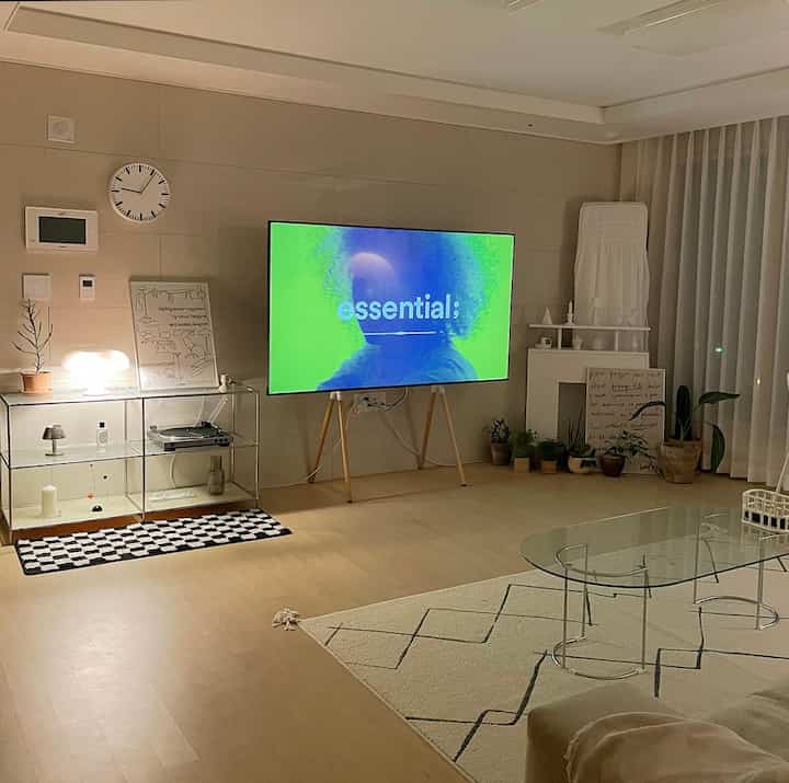 Modern living room with white and wood tones, featuring a TV, glass coffee table, and various plants in a simple arrangement