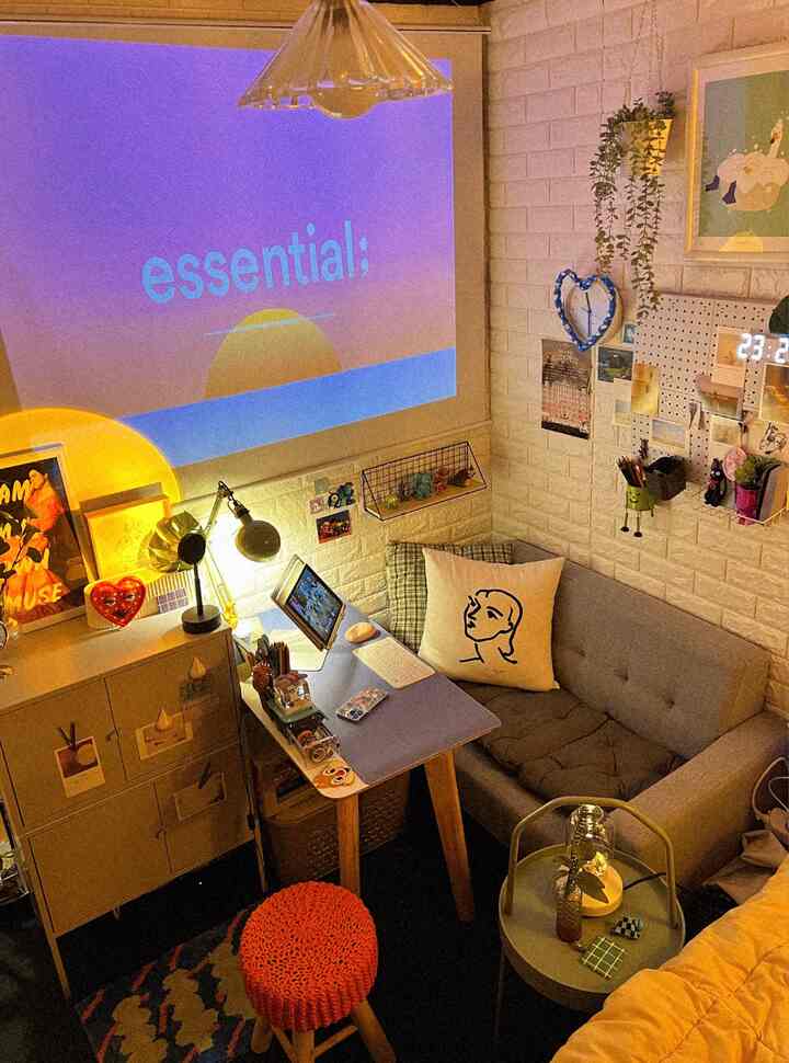 温かみのある色調の小さな作業スペースとリビングルーム、グレーのソファとデスク、壁アート、居心地の良い照明が配置された印象的な空間