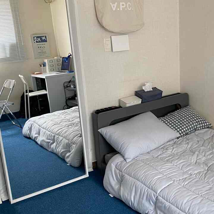 Studio apartment bedroom with blue carpet and pastel-colored bed, featuring a desk and full-length mirror