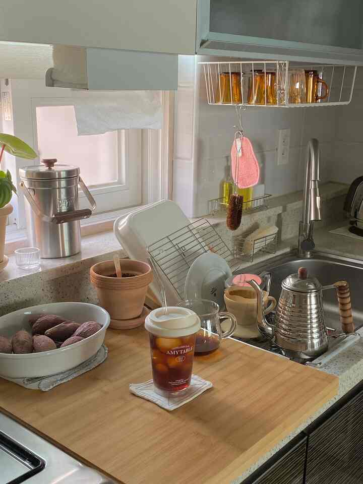 ナチュラルトーンの小さなキッチン、窓際にコーヒー用品と鉢植えがあり、シンク近くに食器乾燥棚が配置された居心地の良いインテリア