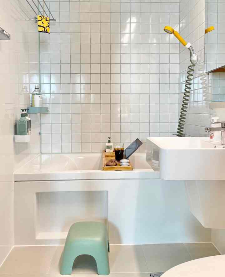 Clean bathroom with white tiled walls, bathtub, yellow showerhead, and light green stool arranged neatly