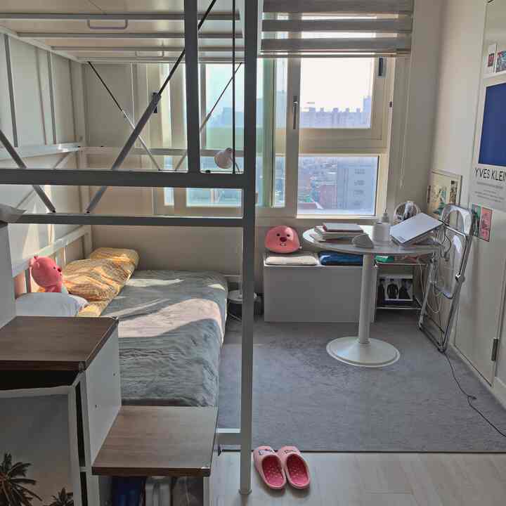 白を基調とした明るいワンルーム寝室にピンクのアクセント、収納ベンチと丸い小さなデスクが配置された実用的な空間