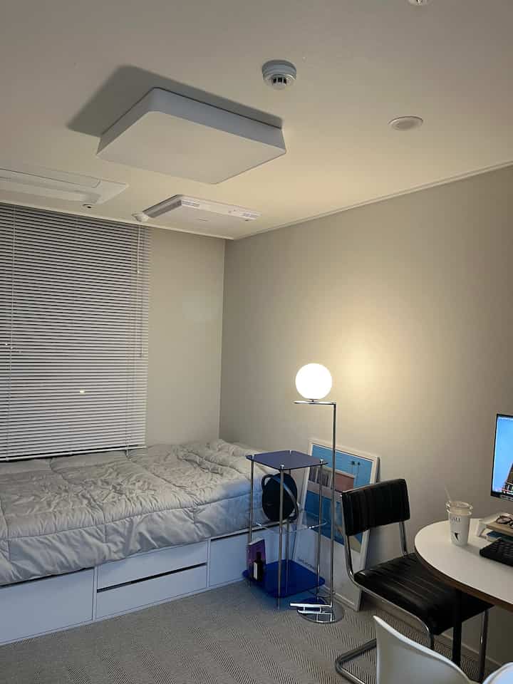 White and gray toned studio apartment featuring single bed, blinds, floor lamp, desk and office chair in a neat space