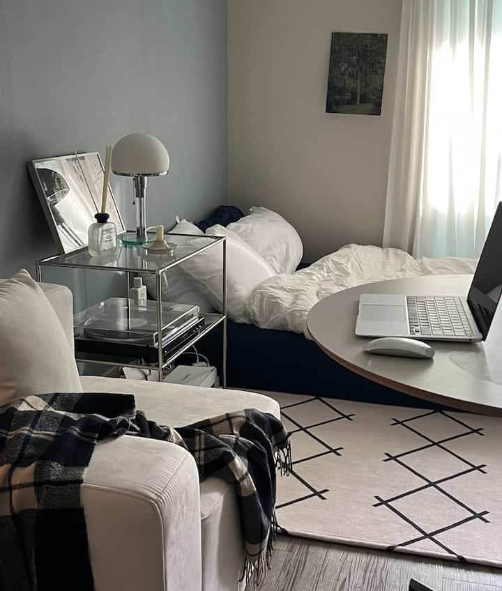 White and gray toned studio bedroom-living room featuring a cozy sofa, bed, and work table with laptop, creating a comfortable space