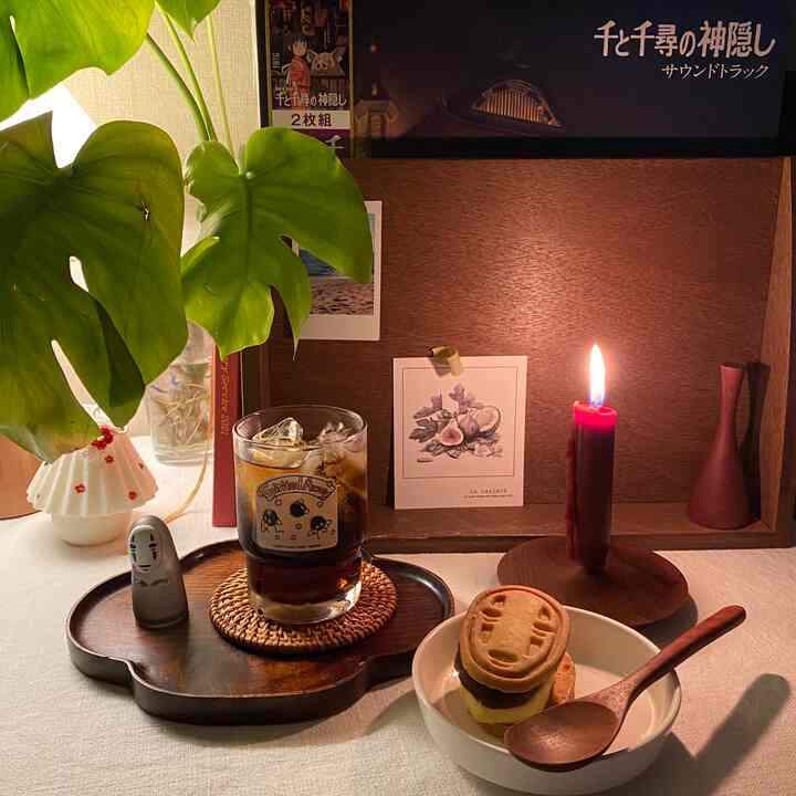 Natural brown-toned home cafe space featuring wooden grain backdrop, monstera plant, No-Face figurine, candle, and iced coffee for cozy atmosphere