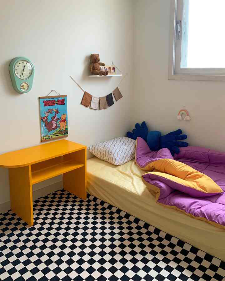 ベージュの壁と白い窓がある子供部屋、黄色の机と紫・黄色の寝具が置かれたベッド、黒白のチェッカーレッグが敷かれた温かみのある空間