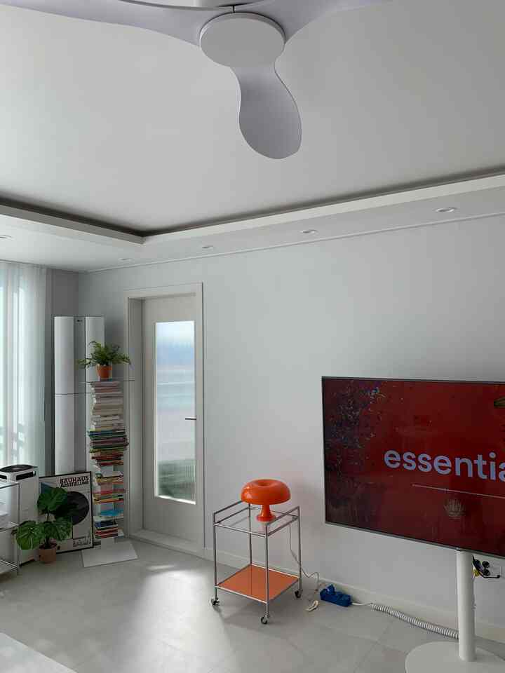 White and orange living room featuring ceiling fan, tall bookshelf, TV, and kitchen cart, creating a modern and natural atmosphere