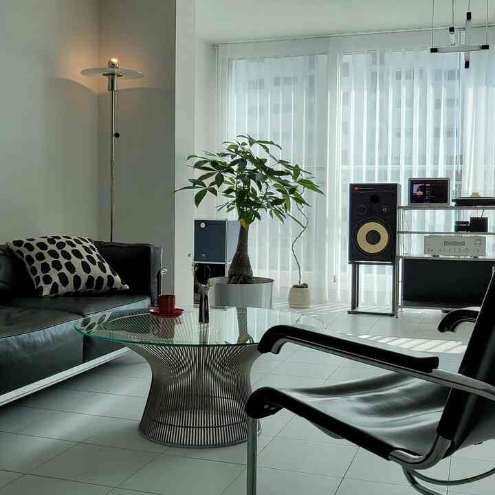 A modern living room with white tiled floor and curtains, featuring a central glass coffee table, black sofa and armchair, speakers, and a floor lamp