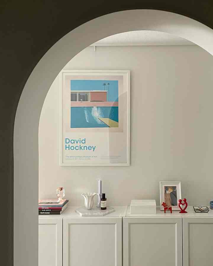 White-toned living room viewed through an archway, featuring a David Hockney poster and small decorative items on a white cabinet in a simple interior