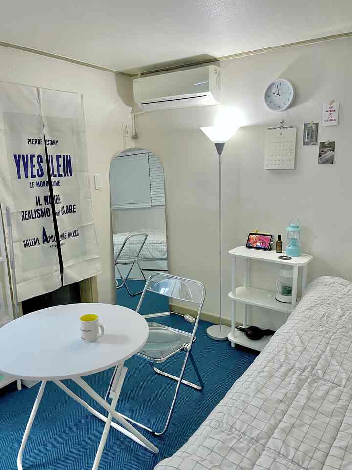 ブルーのラグとホワイトトーンのワンルーム、折りたたみテーブルと透明な椅子が配置された清潔感のあるモダンな5.5坪の空間