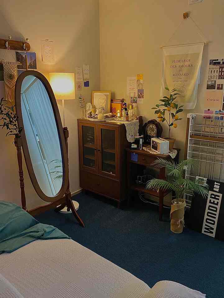 ウッドトーンの家具とブルーカーペットがある居心地の良い一人暮らしの寝室、フロアランプとヴィンテージ時計が配置された自然な雰囲気のインテリア