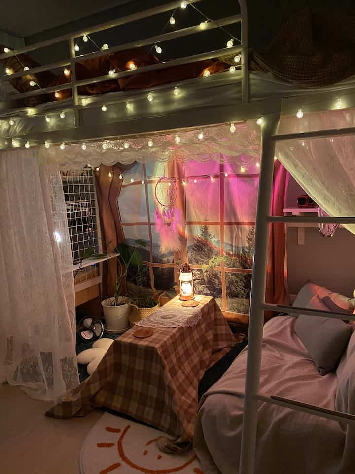 Cozy bedroom with warm lighting featuring a white loft bed, grey sofa, checkered tablecloth, and nature-themed fabric poster