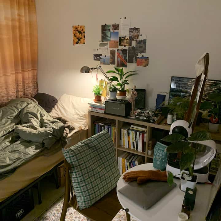 A cozy small bedroom with warm wood tones and beige lighting, featuring a comfortable bed, bookshelf, and round desk