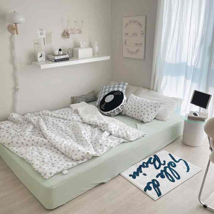 A white and natural tone bedroom featuring a central bed with cushions, wall shelf, and soft natural lighting creating a cozy atmosphere