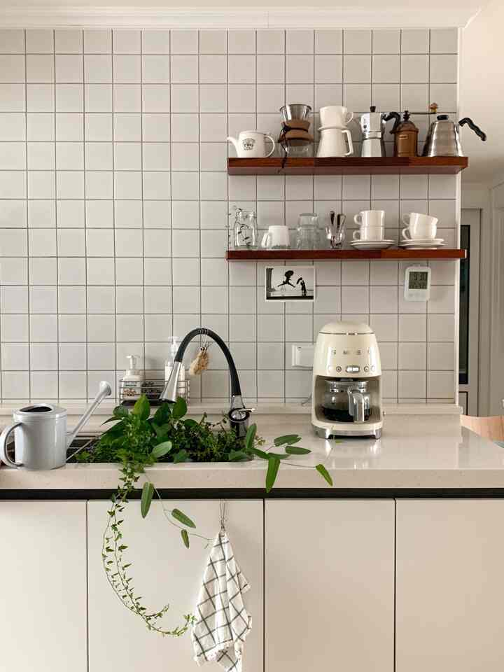 Kitchen with white tiled walls and wooden shelves, green plants growing around the sink, and coffee equipment neatly arranged
