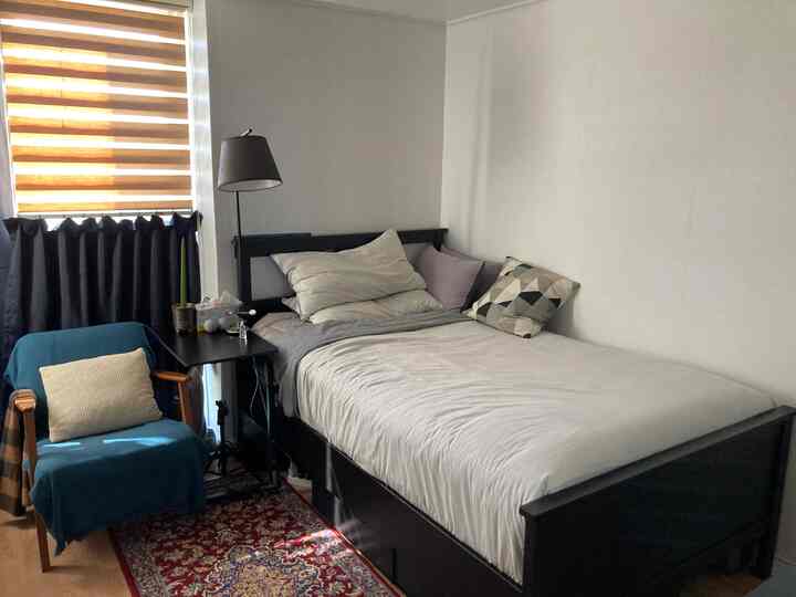 A compact studio bedroom with white walls, black bed frame, blackout curtain tied over blinds, featuring a blue armchair and patterned rug