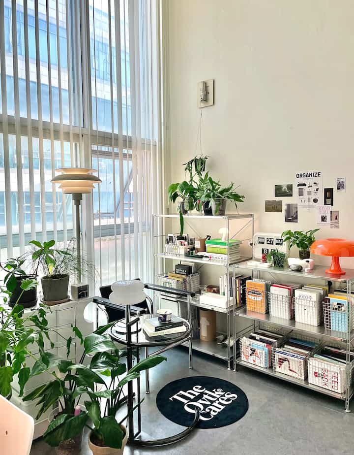 Bright white-walled studio apartment featuring silver metal shelving, abundant greenery, and an orange lamp creating cozy ambiance