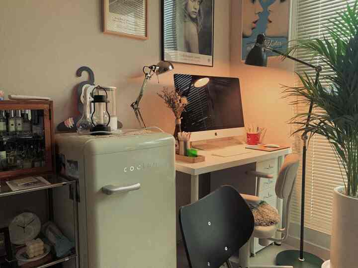 白と木目調が調和した狭いホームオフィス、レトロなミニ冷蔵庫と観葉植物、照明が配置された雰囲気のある空間