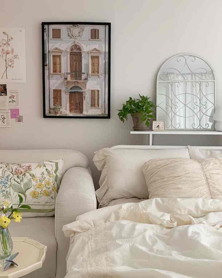 White and ivory toned bedroom space featuring sofa with floral cushion, plant, and decorative wall mirror in a natural modern interior