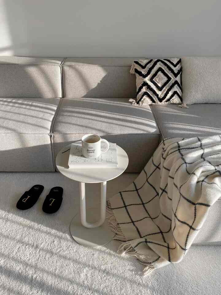 Bright beige-toned living room featuring modular fabric sofa, small round side table, plaid blanket, and cushions arranged for cozy atmosphere