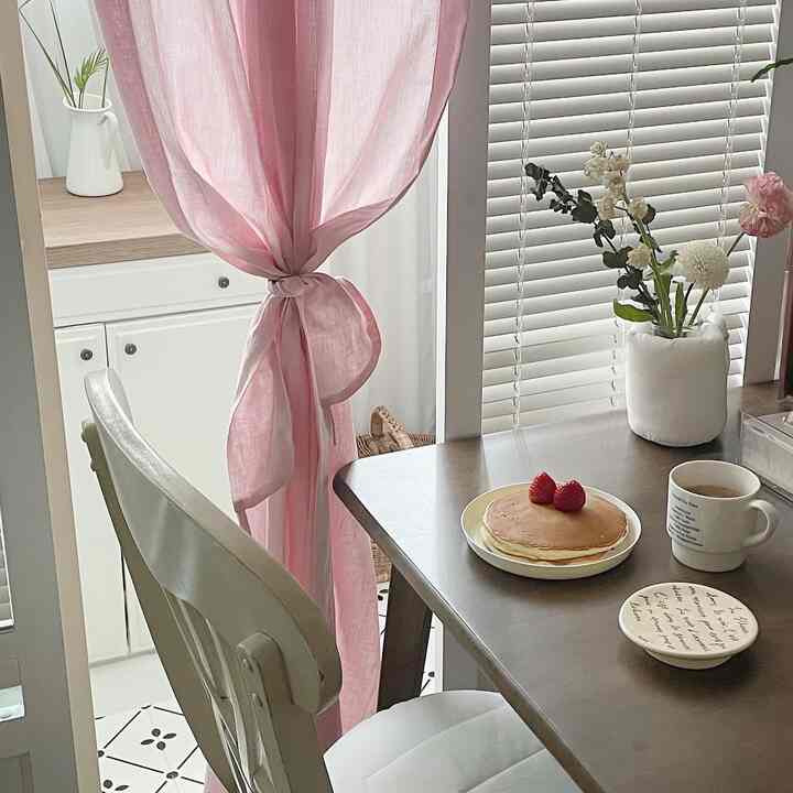 白とピンクの色調のダイニング、食卓にパンケーキとマグカップ、カーテンとブラインドが配置された雰囲気のあるインテリア