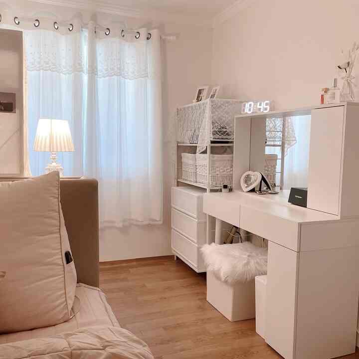A bright white and wood tone space featuring a vanity, storage cabinet, lace curtains, and a floor lamp creating a cozy interior